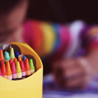 Child wearing a colourful striped jumper colouring in with a box of colouful crayons in the foreground