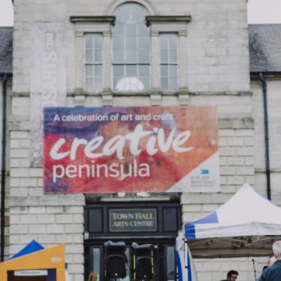 building with a banner saying 'Creative Peninsula' musicians perform in a tent in front of the building with people sitting listening