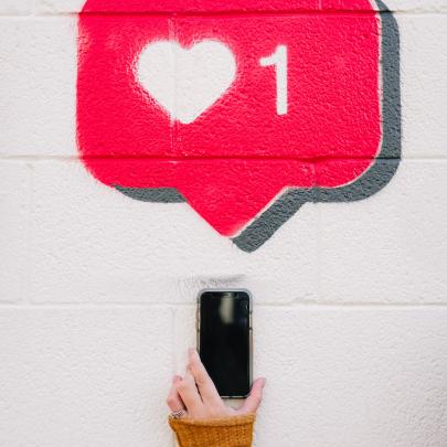 Hand holds a phone up to a brick wall. The wall has graffiti art of a comment box with a heart and the number 1.